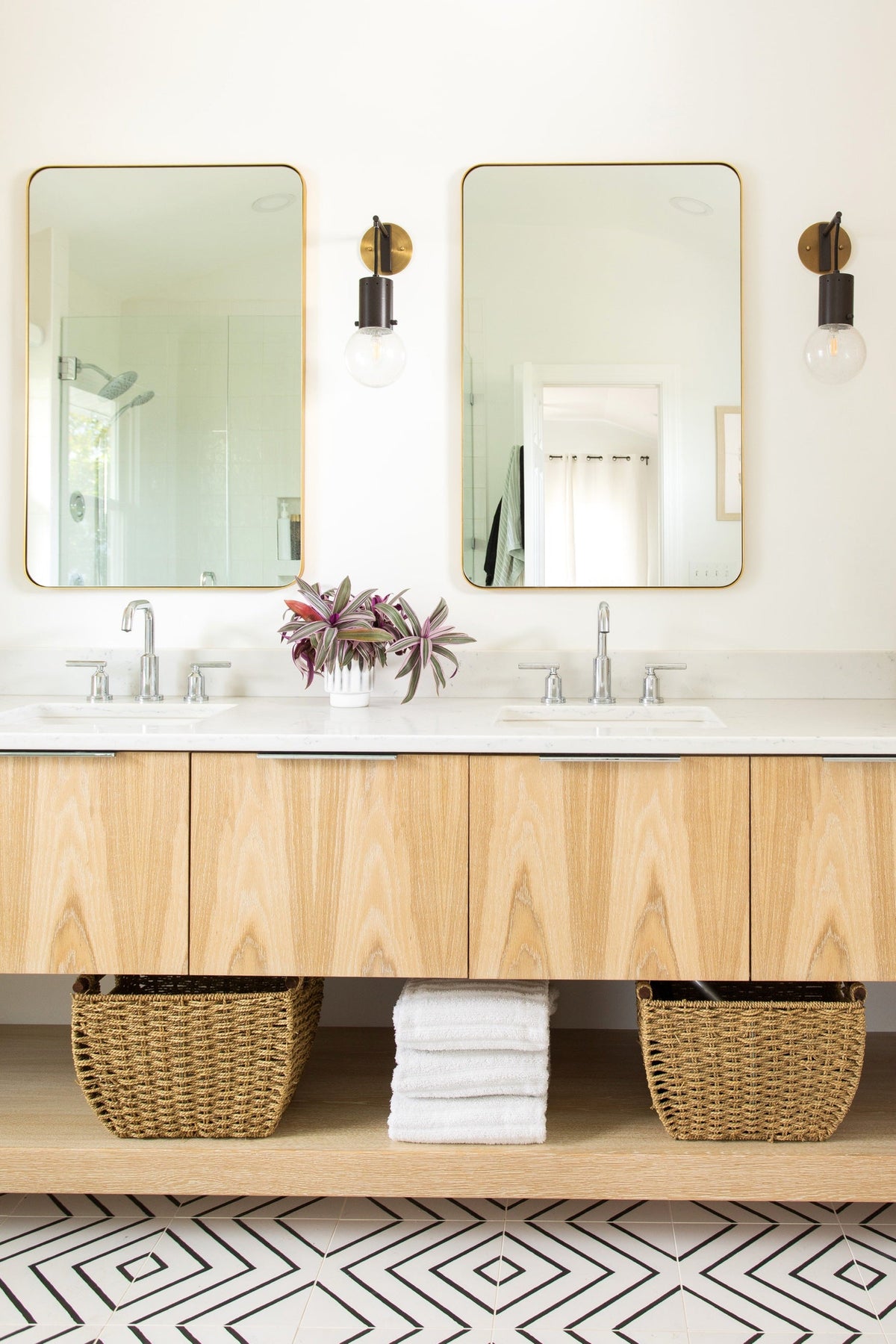 Floating light oak double vanity with two undermount sinks, brass-framed mirrors, and an open shelf holding woven baskets and towels