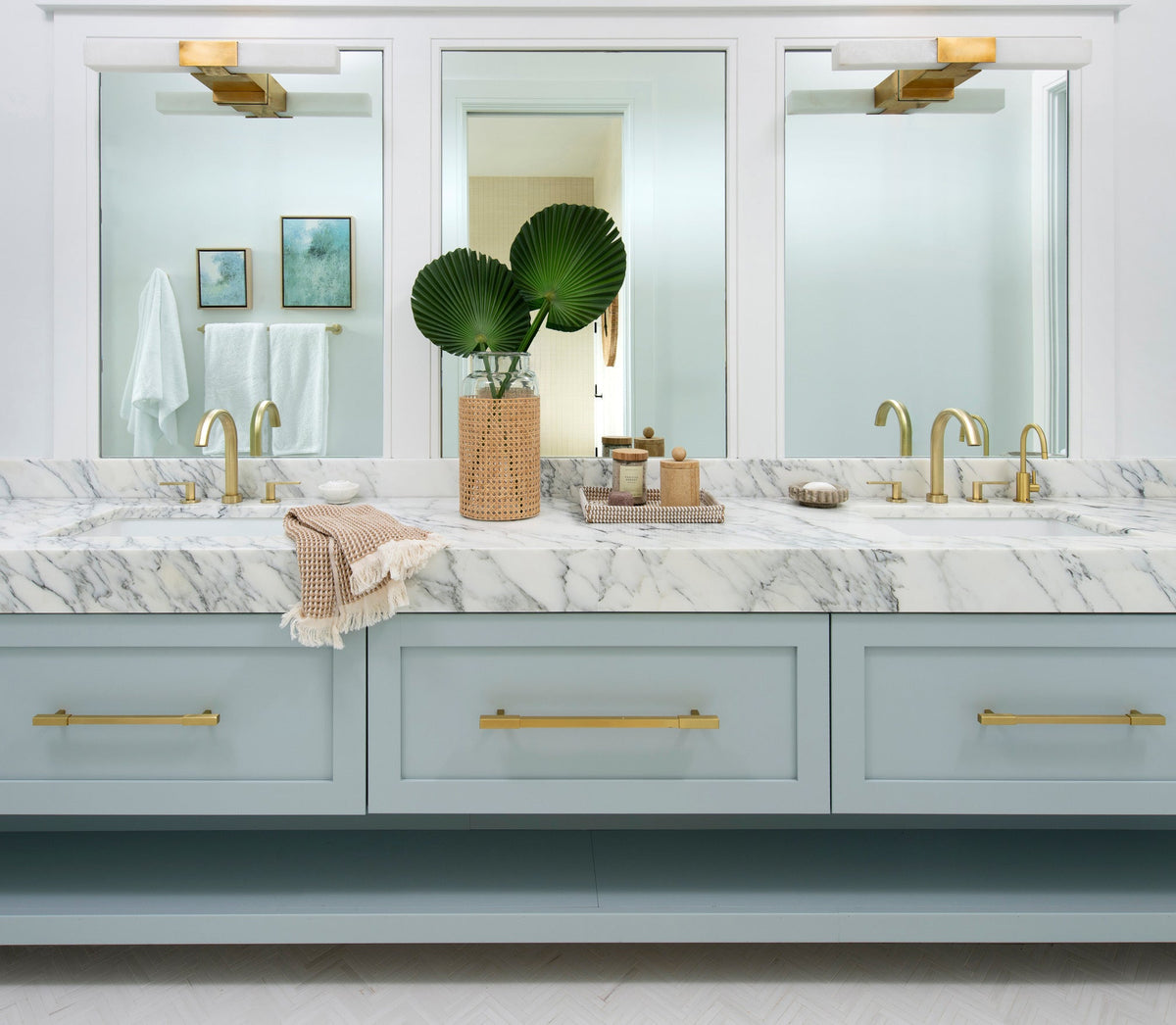 Soft blue double vanity with a marble top, satin brass faucets and pulls, and an open shelf below